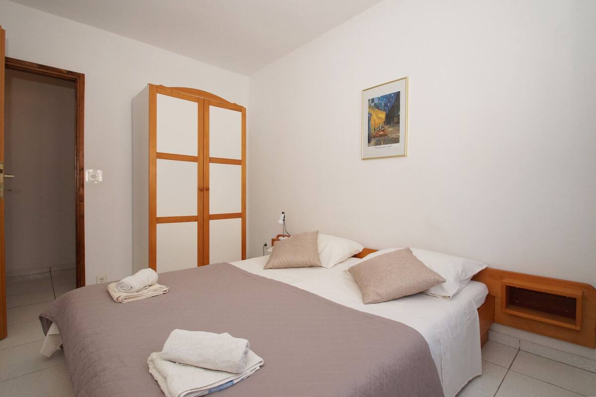 A neatly made bed with a brown bedspread and white pillows, a wooden headboard, a bedside table with a lamp, and a framed picture on the wall.