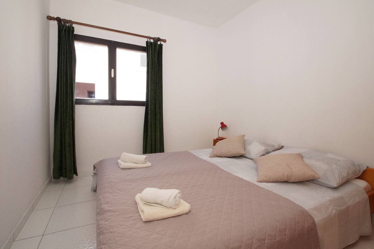 A neatly made bed with a beige bedspread and pillows, a window with green curtains, and a bedside table with a lamp.