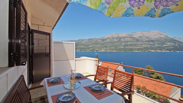 A balcony with a table set for dining, overlooking a mountainous coastline and the sea. - South Island Apartments (Korcula)
