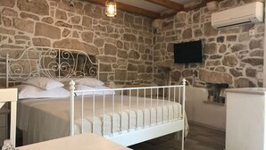 Un dormitorio con pared de piedra, cama blanca, techo de madera y televisor montado.