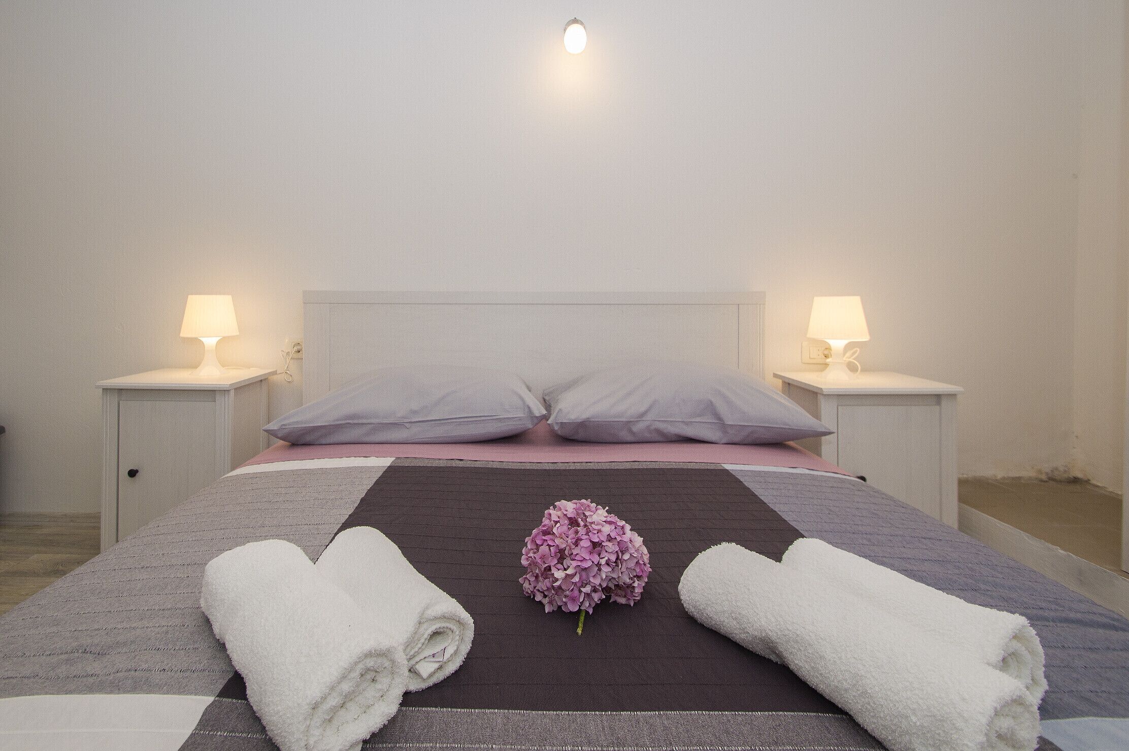 A neatly made bed with a dark brown bedspread, two white pillows, and a pink flower centerpiece. There are two bedside lamps on white nightstands.
