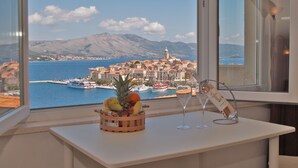 A table with a fruit basket and a wine glass, a bottle of champagne, and a sign, with a view of a coastal town and boats. - Oasis Blue (Korcula)