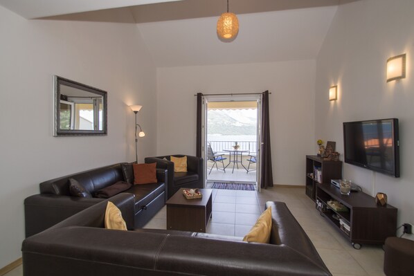 A living room with a sofa, coffee table, and a TV. There is a balcony with a table and chairs. - Oasis Blue (Korcula)