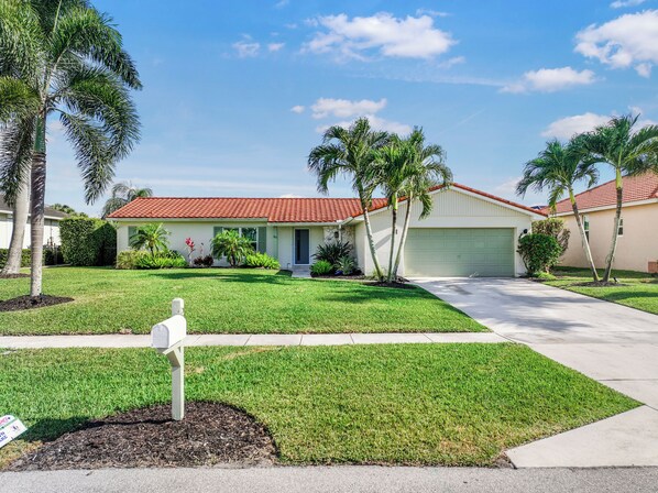 A two-story house with a red-tiled roof, a front yard with grass and palm trees, and a mailbox in the foreground.