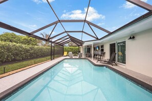 A swimming pool with a glass ceiling, surrounded by a patio with chairs and a table, and a view of a house with a sliding door.