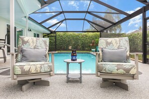 An outdoor patio with two chairs, a table, and a pool in the background. - Nautical home w/ pool, grill & steps to shops (Marco Island)