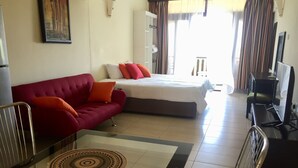 Habitación de hotel con un sofá rojo, una cama con almohadas blancas y anaranjadas, una mesa de vidrio con un mantel individual estampado, un televisor y una silla.