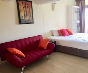 A room with a red sofa, a bed with white and orange pillows, a painting on the wall, and a wardrobe.