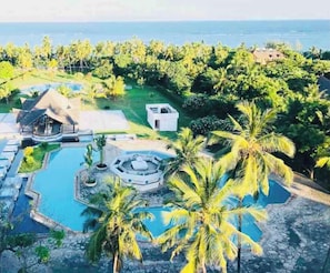 An aerial view of a resort with multiple swimming pools, palm trees, and a view of the ocean.