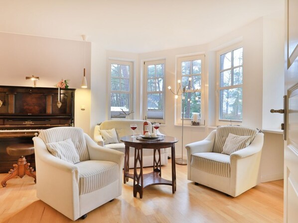 A living room with a piano, two armchairs, a round table, and large windows.