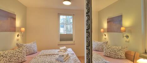 A hotel room with two beds, a window, a mirror, and wall-mounted lamps.