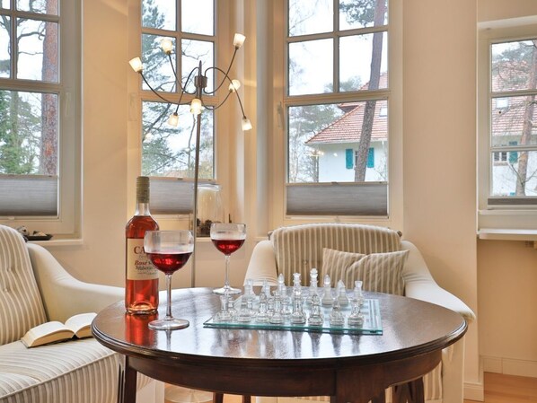 A round table with a chessboard, a bottle of wine, and two glasses of red wine in a room with large windows and striped chairs.