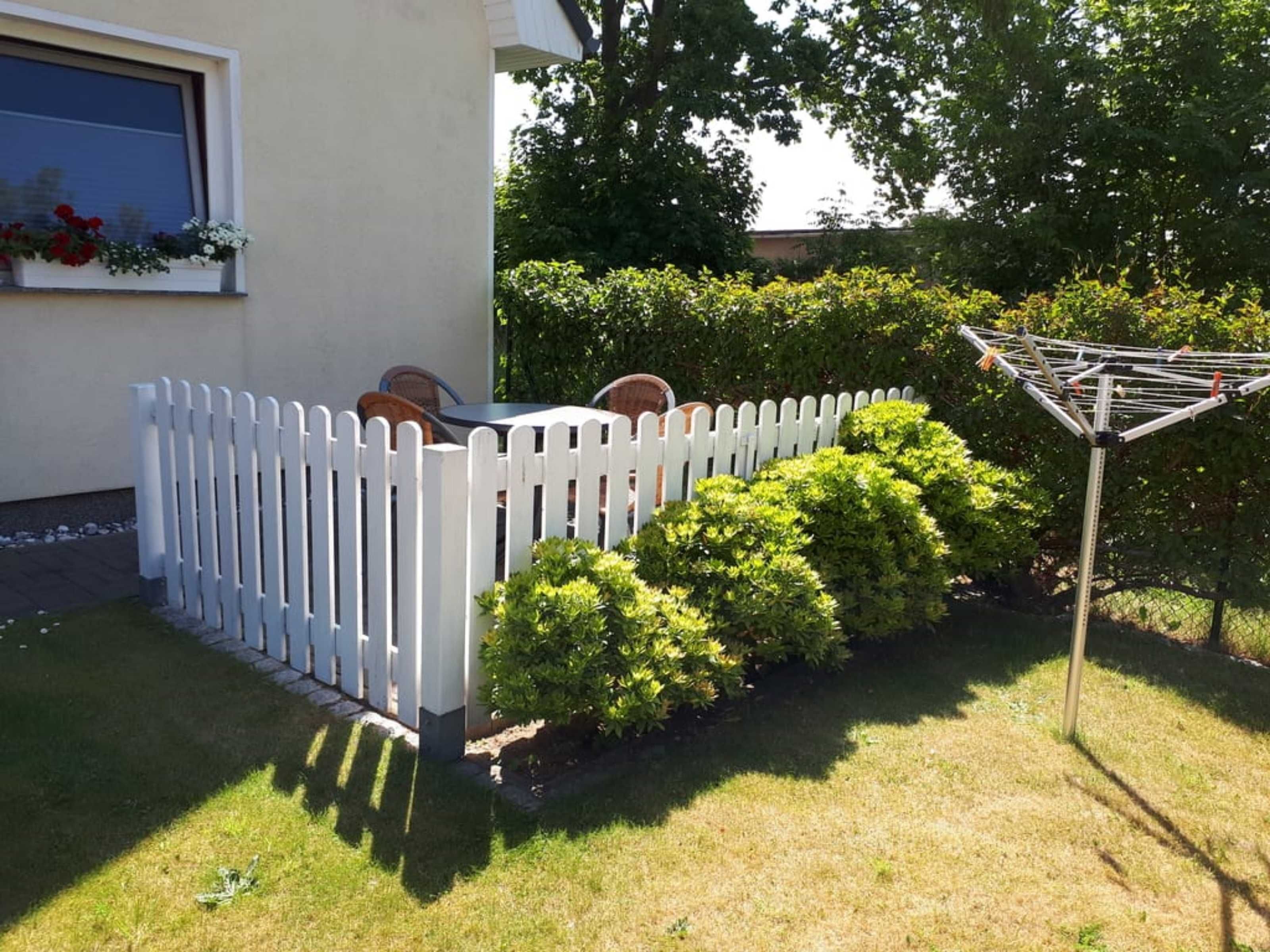 A white picket fence, a small table with chairs, and a clothes drying rack in a garden area.