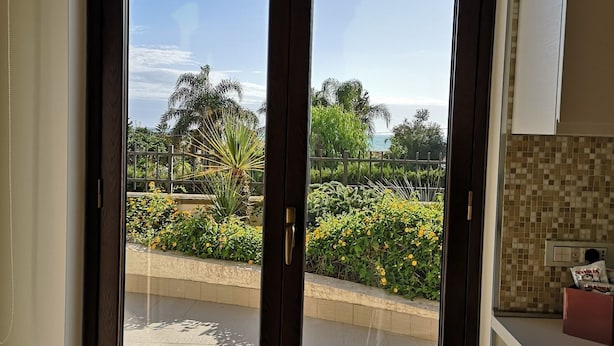 A room with a sliding glass door leading to a balcony with a view of a landscaped garden and a clear sky.
