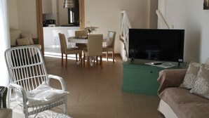 A living room with a sofa, a white wicker chair, a dining table with chairs, a television, and a kitchen area visible through an open door.