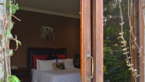 Quarto com cama, mesa de cabeceira, uma obra de arte na parede e vista para um jardim através de uma porta aberta.