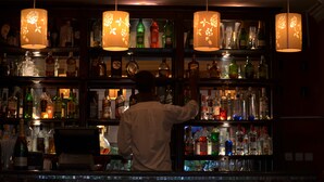 Um balcão de bar com prateleiras repletas de garrafas e copos, e uma pessoa em pé atrás do balcão.