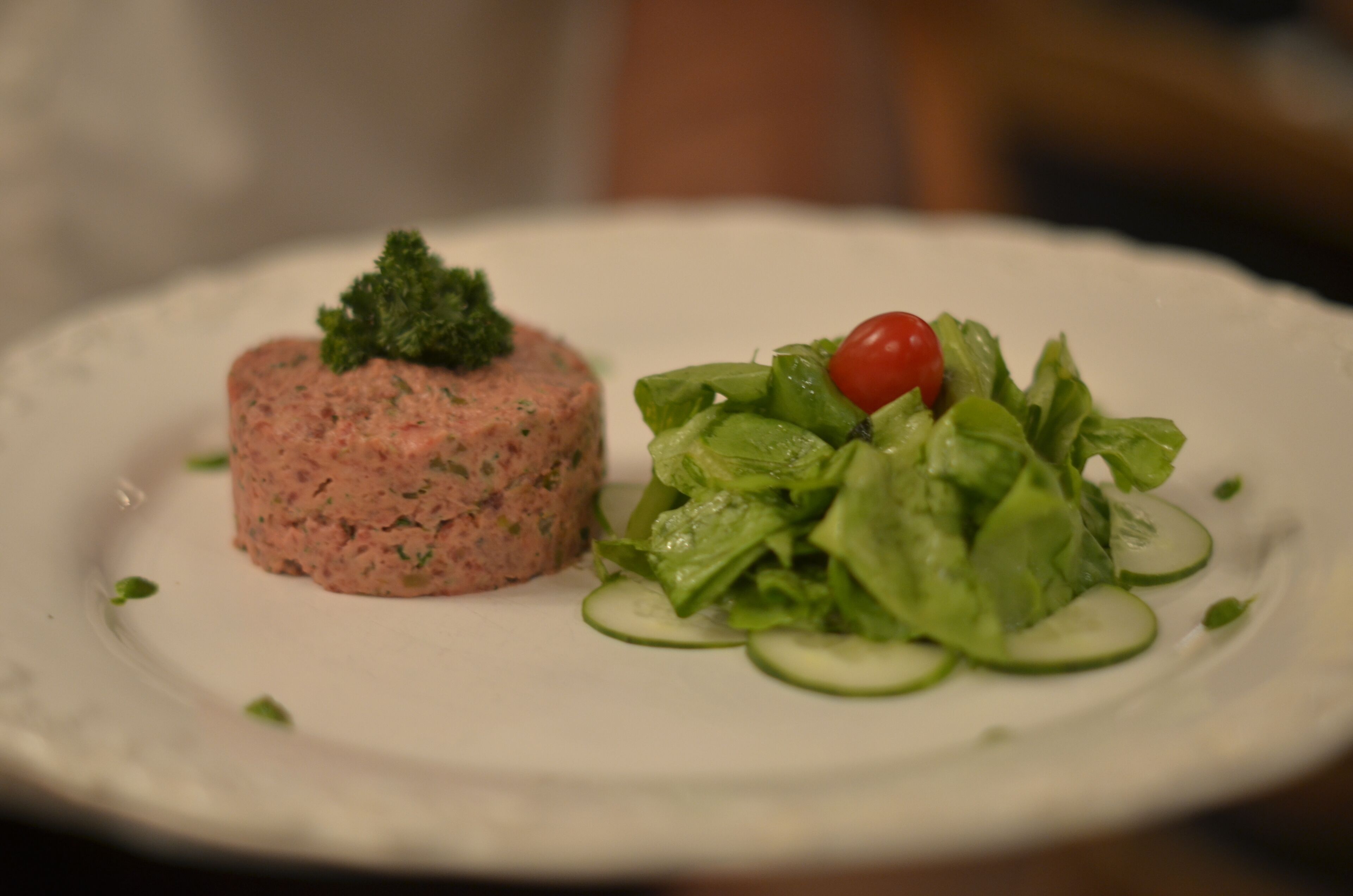 A plate with a pink meat terrine and a green salad with cucumber slices and a cherry tomato.