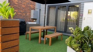 Une terrasse avec une table et des bancs en bois, entourée de gazon synthétique vert, d’un mur en briques et d’une porte-fenêtre coulissante.