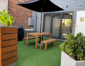 A patio with a wooden table and benches, surrounded by green artificial turf, a brick wall, and a sliding glass door.