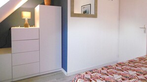 Un dormitorio con techo inclinado, una mesita de noche con lámpara, un aparador, una colcha estampada, un espejo y una decoración con motivos de peces.