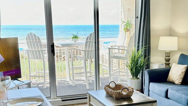 A room with a view of the ocean, a balcony with white chairs, a table with a basket, a sofa, and a lamp.