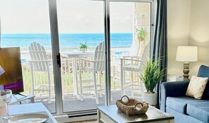 Ett rum med utsikt över havet, en balkong med vita stolar, ett bord med en korg, en soffa och en lampa.