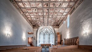 A church interior with wooden pews, a vaulted ceiling with detailed artwork, and arched windows.