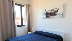 Ein Schlafzimmer mit Bett, Nachttisch, Lampe, einem Fenster mit Vorhängen und einem Bild an der Wand.