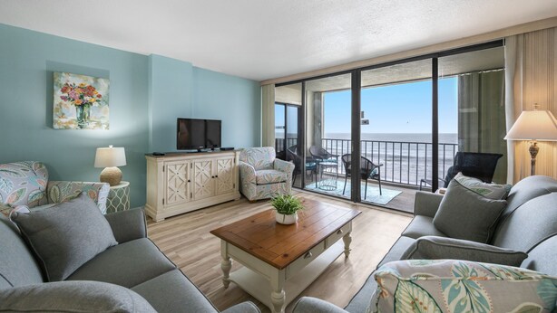 Ein Wohnzimmer mit einer Couch, einem Couchtisch mit Pflanze, einem Fernseher auf einem Schrank und einem Balkon mit Meerblick.