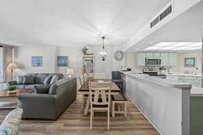 A modern living room with a grey sofa, wooden dining table, and chairs. A kitchen with green cabinets and a microwave is visible.