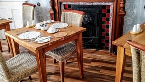 Un coin repas avec une table en bois dressée pour quatre personnes, entourée de chaises en osier. Une cheminée avec un manteau carrelé se trouve au fond de la pièce.