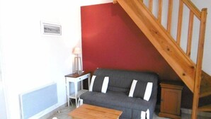Una sala pequeña con un sofá gris, una mesa de centro de madera, una silla blanca, una mesita auxiliar blanca y una escalera de madera.