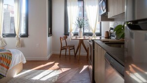 A compact room with a bed, a small dining area with a table and chairs, a kitchen with a refrigerator, and large windows with curtains. - Billy Penn Studio at The Kestrel (Philadelphia)