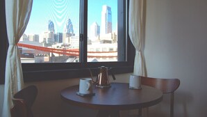 A room with a table, two chairs, a window with curtains, and a view of a cityscape with buildings and a bridge. - Billy Penn Studio at The Kestrel (Philadelphia)