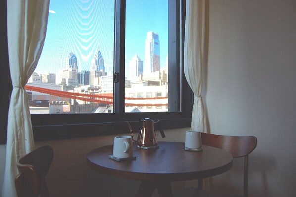 A room with a table, two chairs, a window with curtains, and a view of a cityscape with buildings and a bridge.