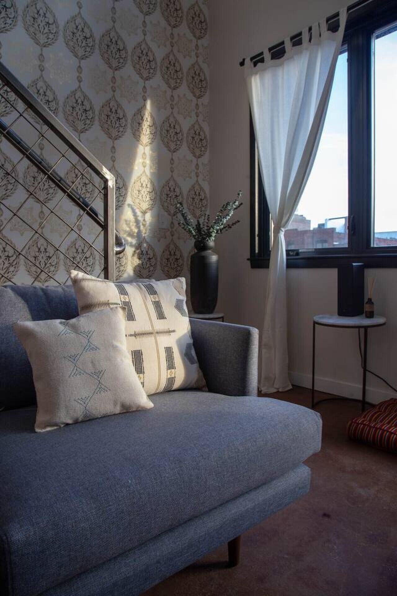 A modern living room with a grey sofa, patterned cushions, a potted plant, and a view of the city through a window with white curtains.
