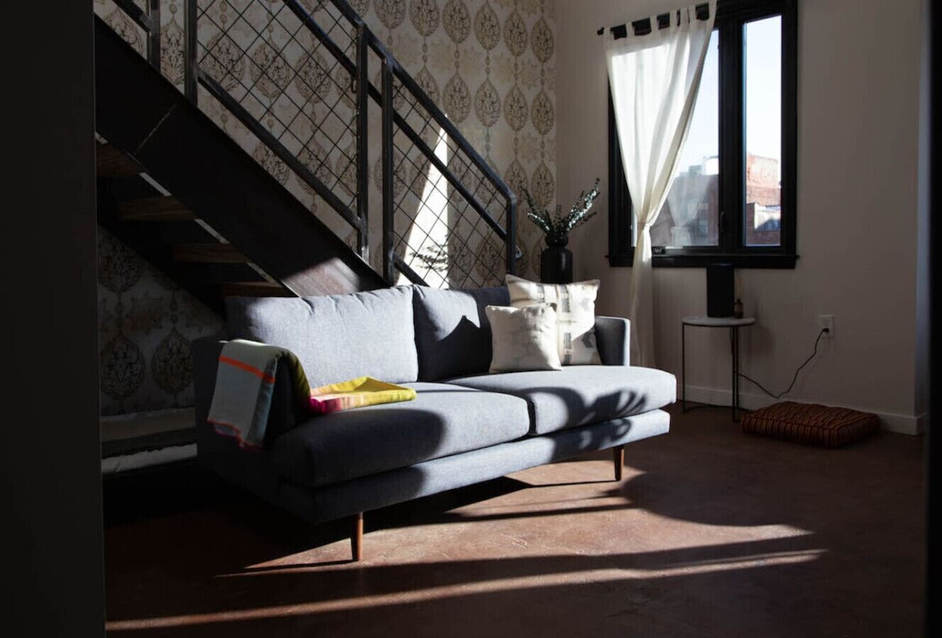 Ett modernt vardagsrum med en grå soffa, en trappa, ett fönster med gardiner och ett litet sidobord.