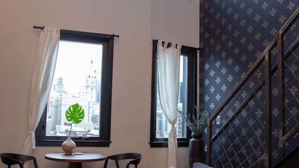 A living room with a gray sofa, two chairs, a small table with a plant, and a city view through large windows. - Hive Room Loft at The Kestrel (Philadelphia)