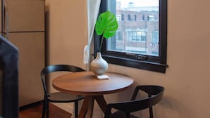 A small dining area with a round table, two chairs, a white vase with a green plant, and a window with a curtain. - Hive Room Loft at The Kestrel (Philadelphia)