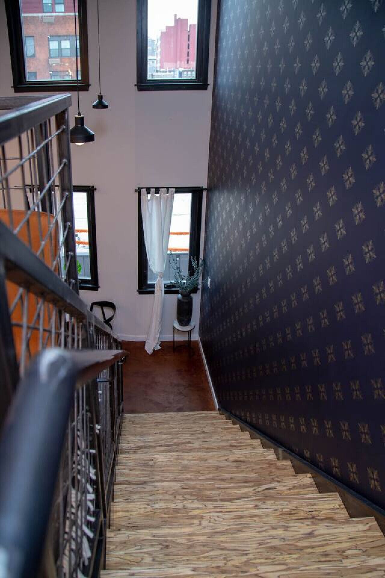 Una scala con pavimento in legno, ringhiera nera e vista all'esterno attraverso una finestra con tende bianche.
