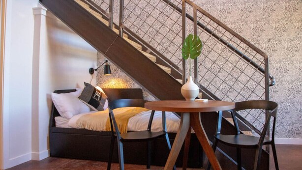 A cozy bedroom with a bed, a small table, and a chair. There is a staircase with a metal railing and a decorative plant. - Mayor`s Study Loft at The Kestrel (Philadelphia)