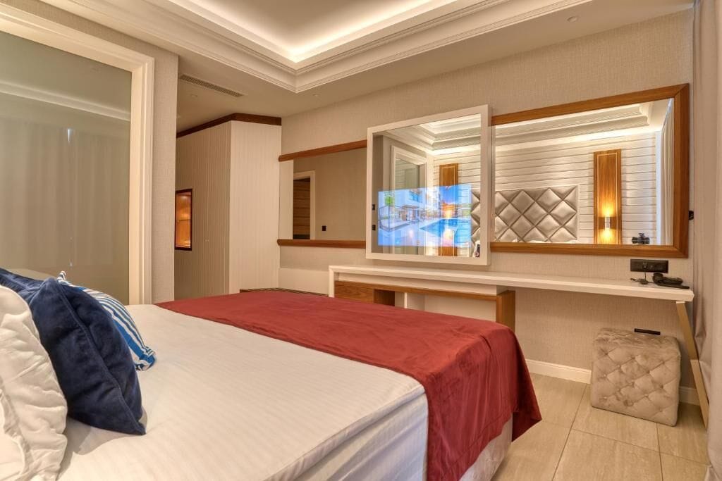 Une chambre d’hôtel avec un lit, un grand miroir, une table de chevet et un petit pouf.