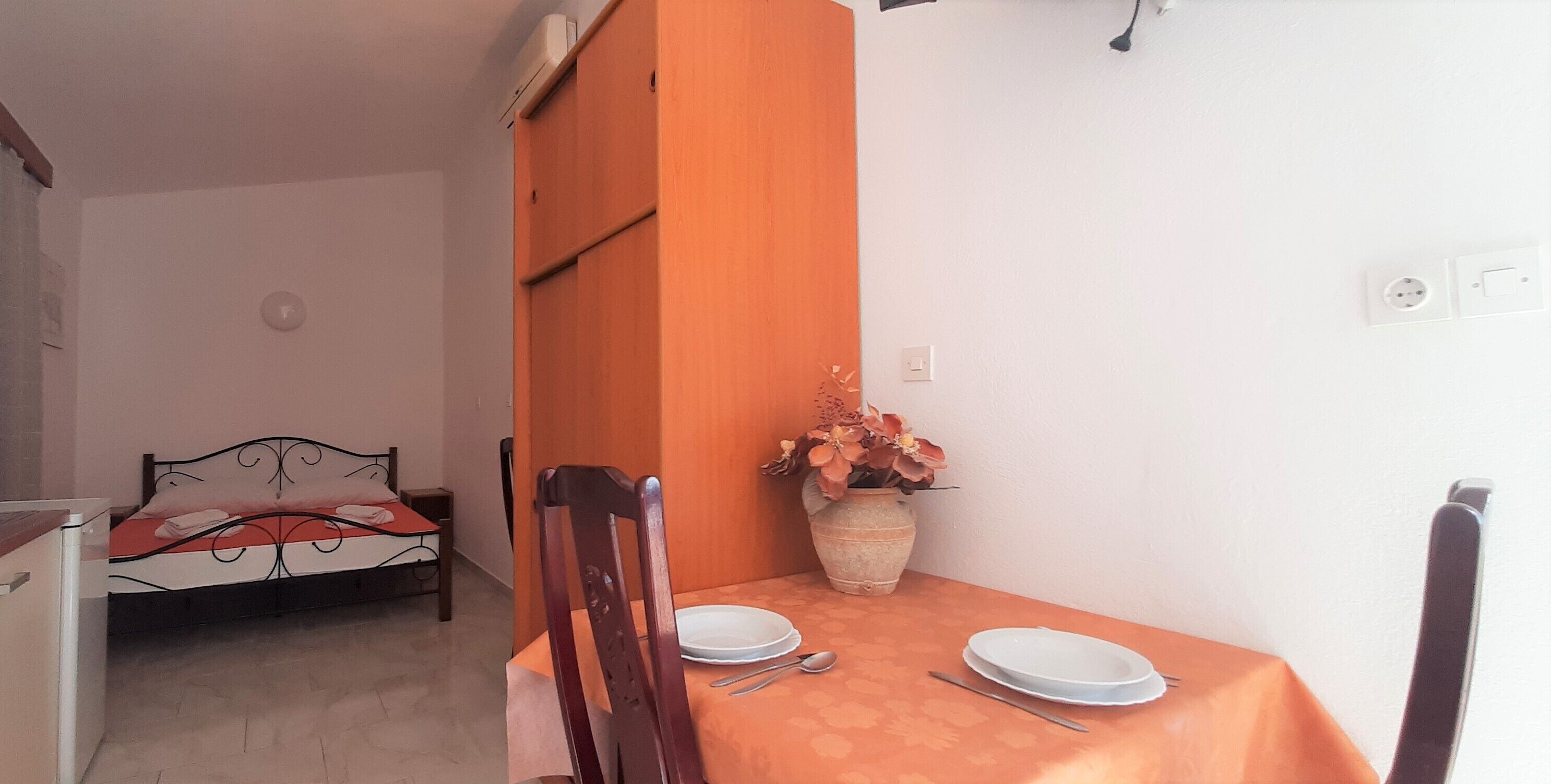 A compact room with a bed, a dining table with plates and cutlery, and a potted plant.
