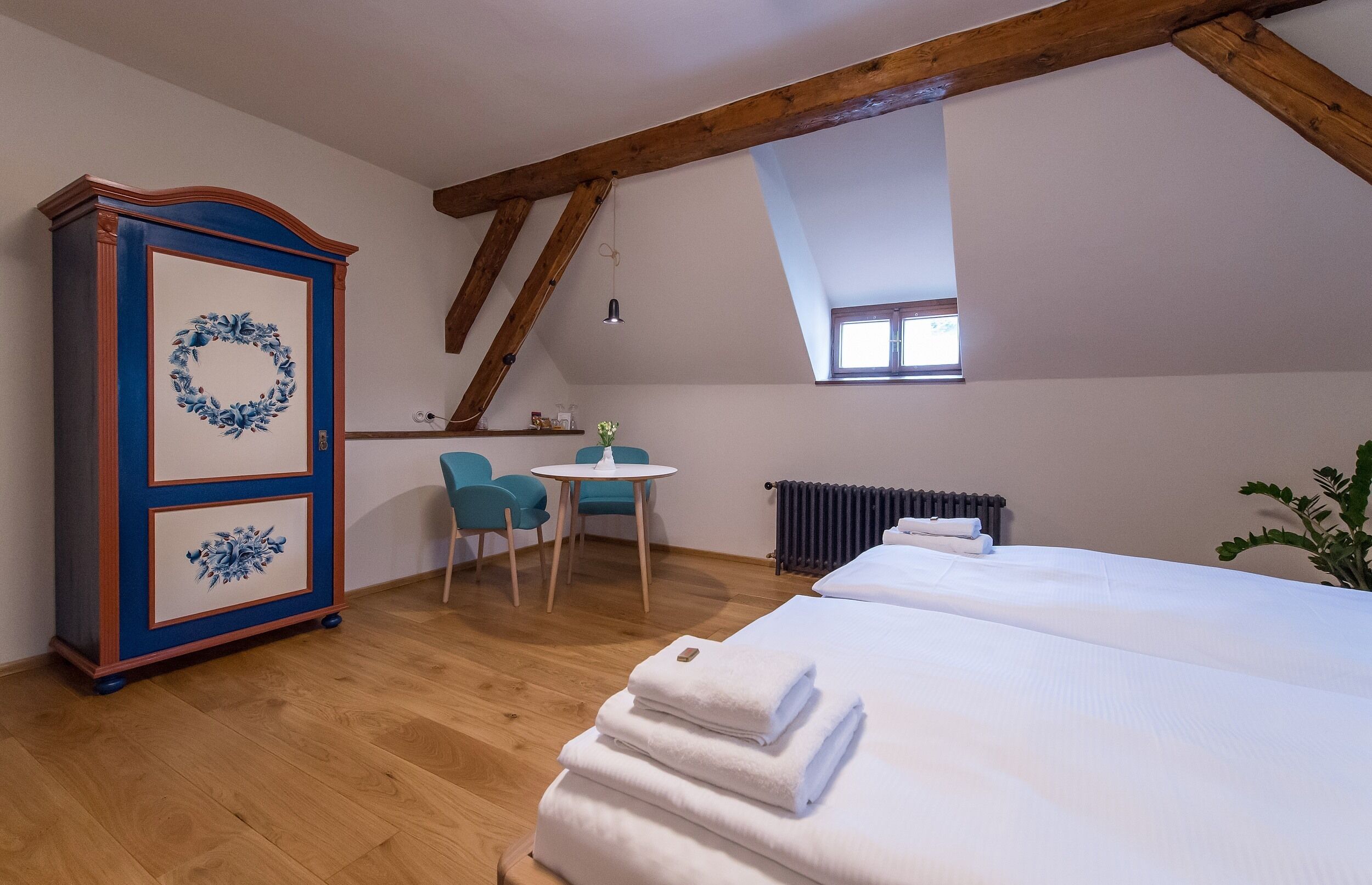 A bedroom with a wooden ceiling, a bed with white linens, a blue and white cabinet, a small round table with chairs, and a window with a view.