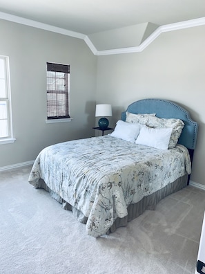 A bedroom with a bed, a nightstand, a lamp, a window with blinds, and a carpeted floor.