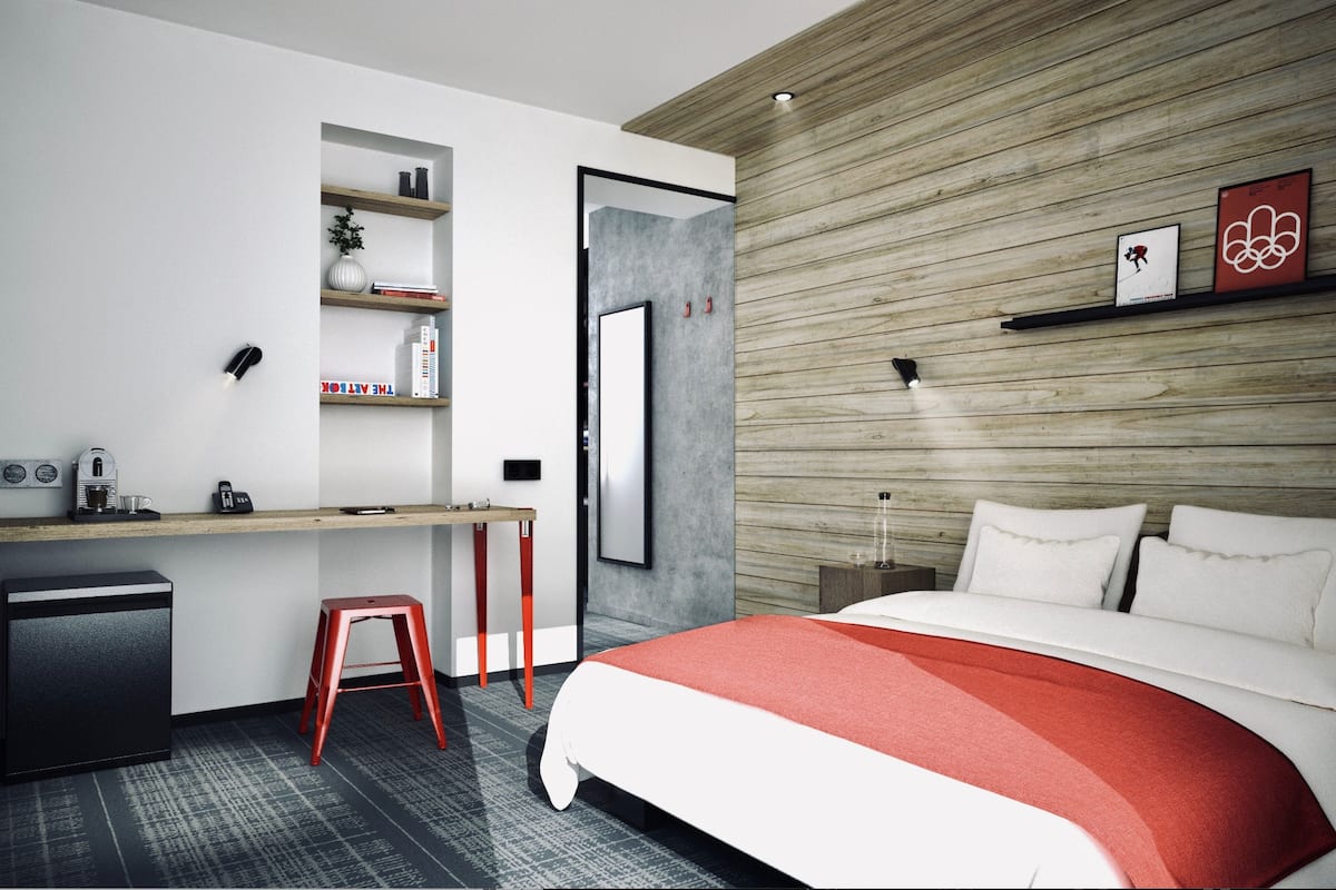 Habitación de hotel moderna con cama, escritorio, silla y un taburete pequeño. Se aprecia una estantería con libros y un jarrón en la pared.
