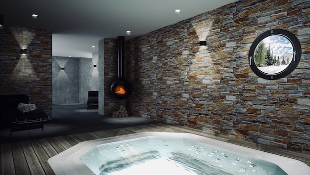 A modern spa room with a stone wall, a circular window, a fireplace, and a hot tub.