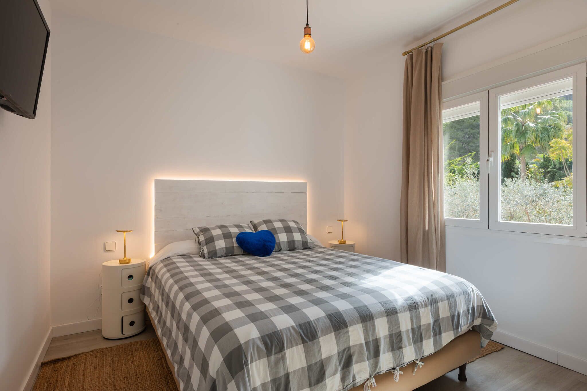 Ett sovrum med ett rutigt sängöverkast, en blå kudde, sänglampor och ett fönster med utsikt över grönskan.