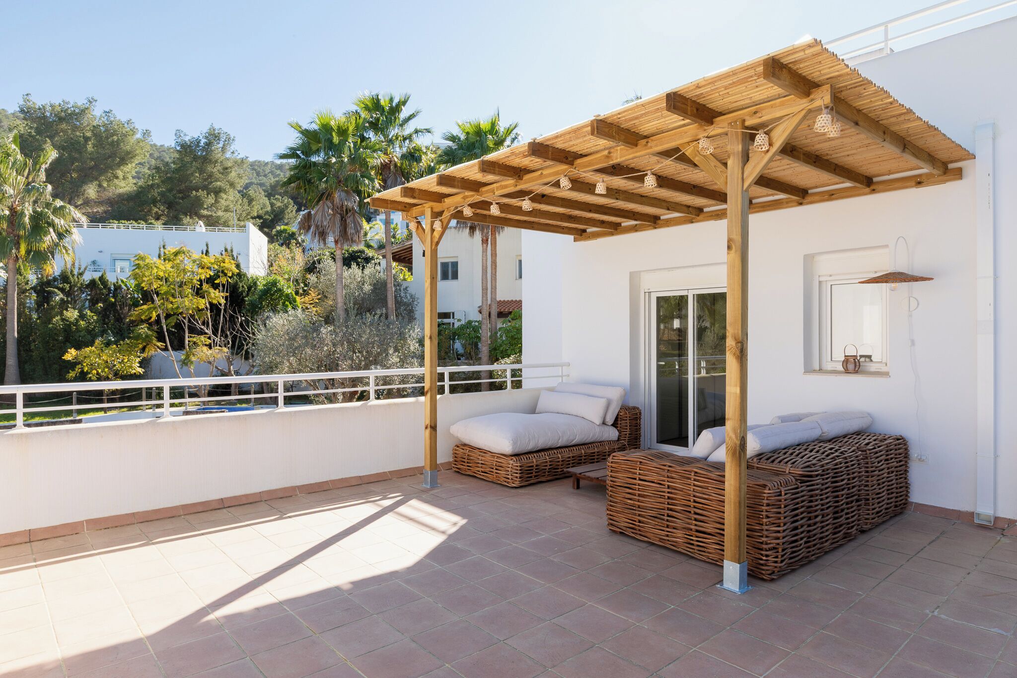 A rooftop terrace with wicker furniture, a pergola, and a view of palm trees and buildings.